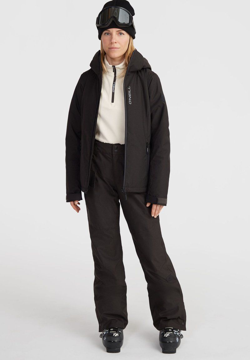 Schwarze Skijacke mit Kapuze, Reißverschluss und Logodetails, kombiniert mit schwarzen Skihosen und Stiefeln. Outfit für den Wintersport entworfen.