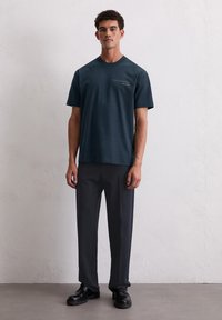 Dunkelblaues Baumwoll-T-Shirt mit kurzen Ärmeln und Rundhals-Ausschnitt, mit schwachem Text auf der linken Seite. Kombiniert mit dunkelgrauen Hosen und schwarzen Schuhen.