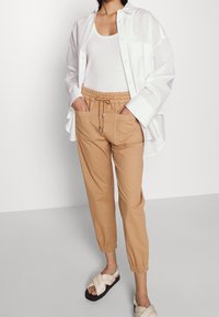 Khaki tapered pants with elastic waistband, side pockets, and drawstrings paired with a white tank top and loose white shirt. Sandals featured.