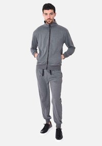 Felpa grigia con zip, colletto alto e tasche, abbinata a pantaloni della tuta tapered coordinati e sneakers nere con suola bianca.
