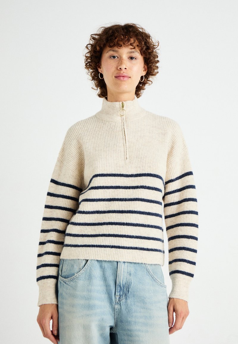 Cream and navy striped sweater with a high collar, gold zipper, and ribbed texture. Styled with light blue denim pants.