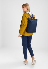 Mochila de tela azul marino con asas superiores y correas para los hombros, contra un fondo claro, llevada con un chubasquero amarillo y jeans azules.