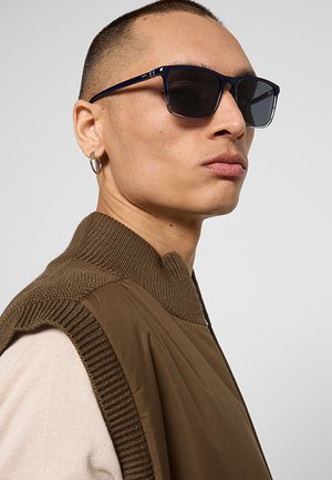 Uomo con capelli corti che indossa occhiali da sole neri rettangolari, un gilet marrone di maglia sopra una camicia chiara e un orecchino a cerchio nell’orecchio sinistro.