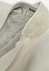 Veste de couleur claire avec un tissu extérieur texturé. L'intérieur présente une doublure à motif géométrique et un col en feutre doux.