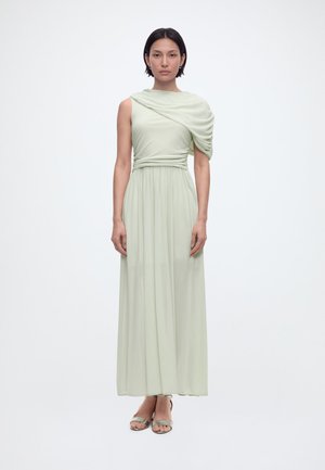 Kvinne stående, iført en lys grønn, lang kjole med én skulder og rynkedetaljer, samt matchende åpne sandaler på en enkel bakgrunn.