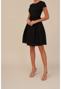 Robe noire à manches courtes, tissu texturé, corsage ajusté et jupe évasée. Associée à des sandales à talons hauts métalliques avec lanières.