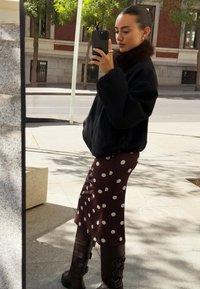 Veste oversized noire avec un col en fourrure, jupe en pois bordeaux, bottes marron jusqu'au genou avec des boucles, et un smartphone à la main.