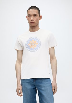 Junger Mann trägt ein weißes T-Shirt mit kurzen Ärmeln und einem bunten, runden Blumenlogo sowie blaue Jeans vor einfarbigem Hintergrund.