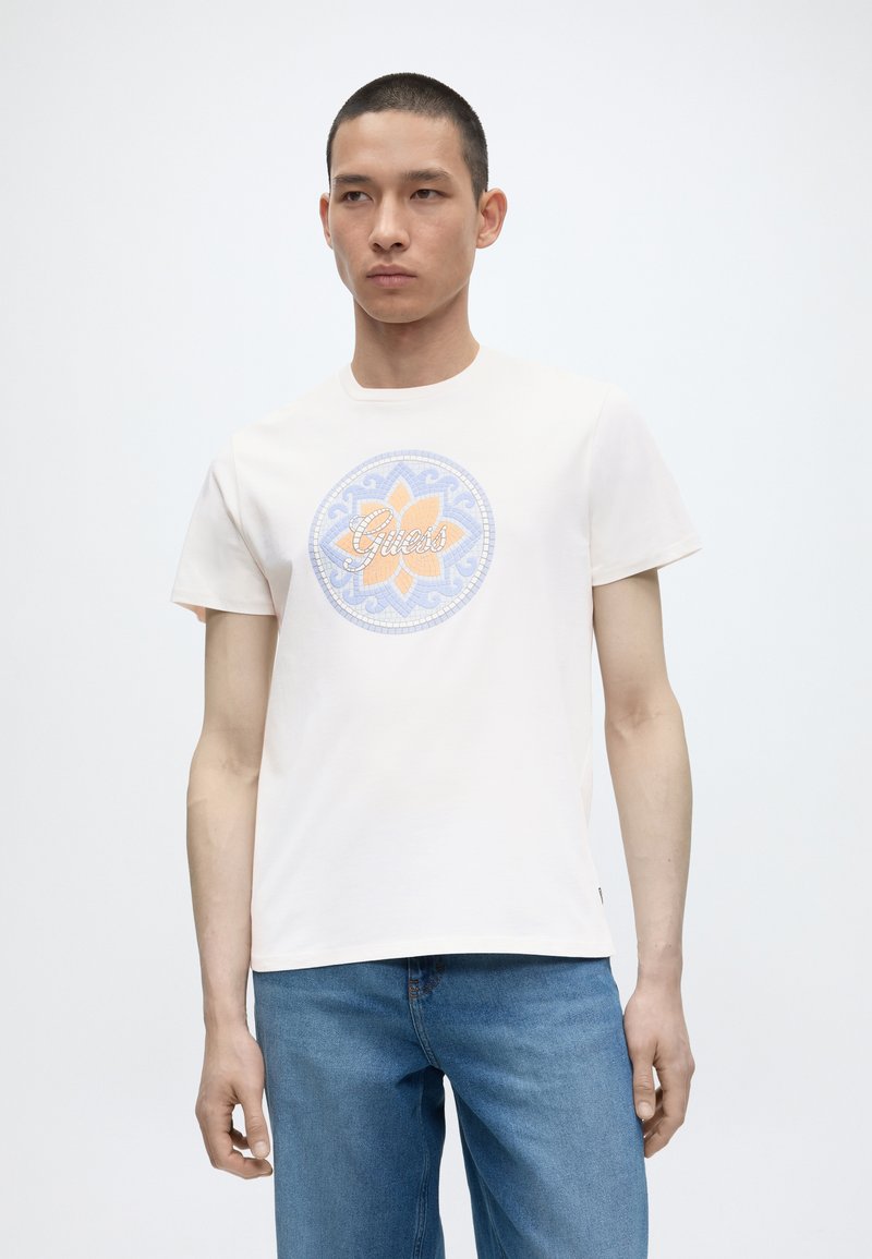 Νέος άνδρας που φοράει λευκό κοντομάνικο T-shirt με πολύχρωμο κυκλικό λογότυπο με λουλούδια και μπλε τζιν σε μονόχρωμο φόντο.