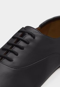 Chaussure Oxford en cuir noir avec laçage fermé et bout arrondi, présentant des coutures détaillées et une texture lisse.