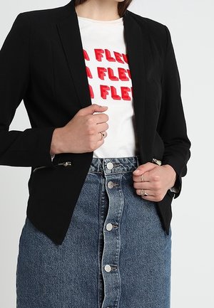 Vrouw draagt een zwarte blazer over een wit t-shirt met rode tekst, gecombineerd met een hoge taille denim rok met knopen aan de voorkant, handen die de blazer aanpassen.