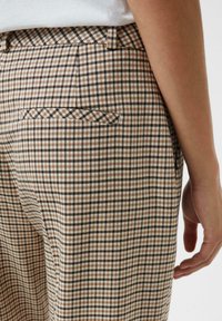 Primo piano di una persona che indossa dei pantaloni a quadri beige e marroni con una tasca welt posteriore e una parte superiore bianca visibile all'altezza della vita.