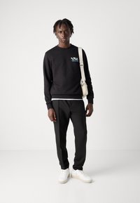 Zwarte sweatshirt met een grafische patch, gecombineerd met zwarte sportbroeken en witte sneakers. De stof is soepel, met een relaxte fit.