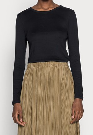 Persoon die een zwart shirt met lange mouwen draagt, ingestopt in een beige geplooide rok met hoge taille, tegen een effen achtergrond.