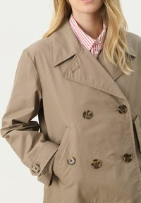 Beige kaksirivinen trench-takki, jossa on lovellinen kaulus, suuret kilpikonnankuoripainikkeet ja sivutaskut, valmistettu sileästä kankaasta.