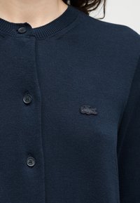 Cardigan en maille bleu marine avec col rond, boutons noirs et petit logo crocodile brodé sur la poitrine gauche.