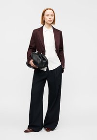 Μπορντό ραμμένο σακάκι πάνω από λευκό πουκάμισο, συνδυασμένο με μαύρο παντελόνι με φαρδιές πιέτες. Κρατώντας μια μαύρη τσάντα-φάκελο, με λεία υφή και στρογγυλεμένο σχήμα.