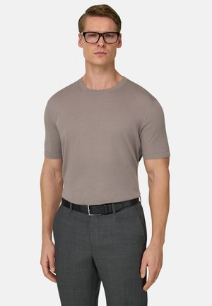 Mann trägt taupefarbenes kurzärmliges Strickhemd, das in graue Anzughose mit schwarzem Gürtel gesteckt ist, trägt eine schwarzgerahmte Brille und steht vor weißem Hintergrund.