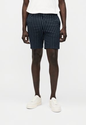 Homme portant un short bleu marine à rayures verticales blanches, associé à des baskets blanches et à une chemise blanche rentrée.