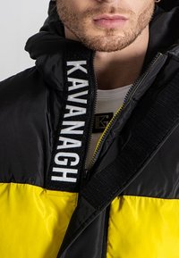 Pufferjakke i sort og gul, med en polstret design, lynlåslukning, logoaccent i hvid og justerbare stropper.