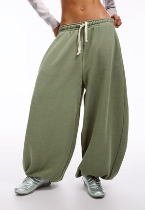 Egy személy laza, olívazöld, széles szárú nadrágot visel, melynek derékán húzózsinór található, valamint ezüst színű tornacipőt, és fehér háttér előtt áll.