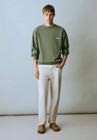 Felpa verde oliva con colletto a girocollo, abbinata a pantaloni beige chiaro e scarpe slip-on marroni. Design semplice e casual con loghi minimi.