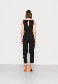 Mono negro acanalado con un diseño de espalda con ojo de llavero y cintura ceñida, combinado con sandalias naranja de tacón. Fondo neutro y plano.