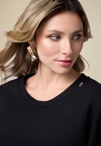 Goldene metallische Ohrringe in abstrakter Form kombiniert mit einem schwarzen Sweatshirt, das über ein dezentes Halsdetail verfügt. Glatte Textur; zeitgemäßes Design.