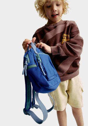 Blauer Rucksack mit grünen Akzenten, mehreren Reißverschlusstaschen und gepolsterten Schulterriemen. Ein Kind hält ihn vor einem schlichten Hintergrund.