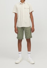 Korte mouwen crèmekleurig shirt met een borstzak, gecombineerd met opgerolde olijfgroene shorts. Witte sneakers en witte sokken met zwarte branding maken de outfit compleet.