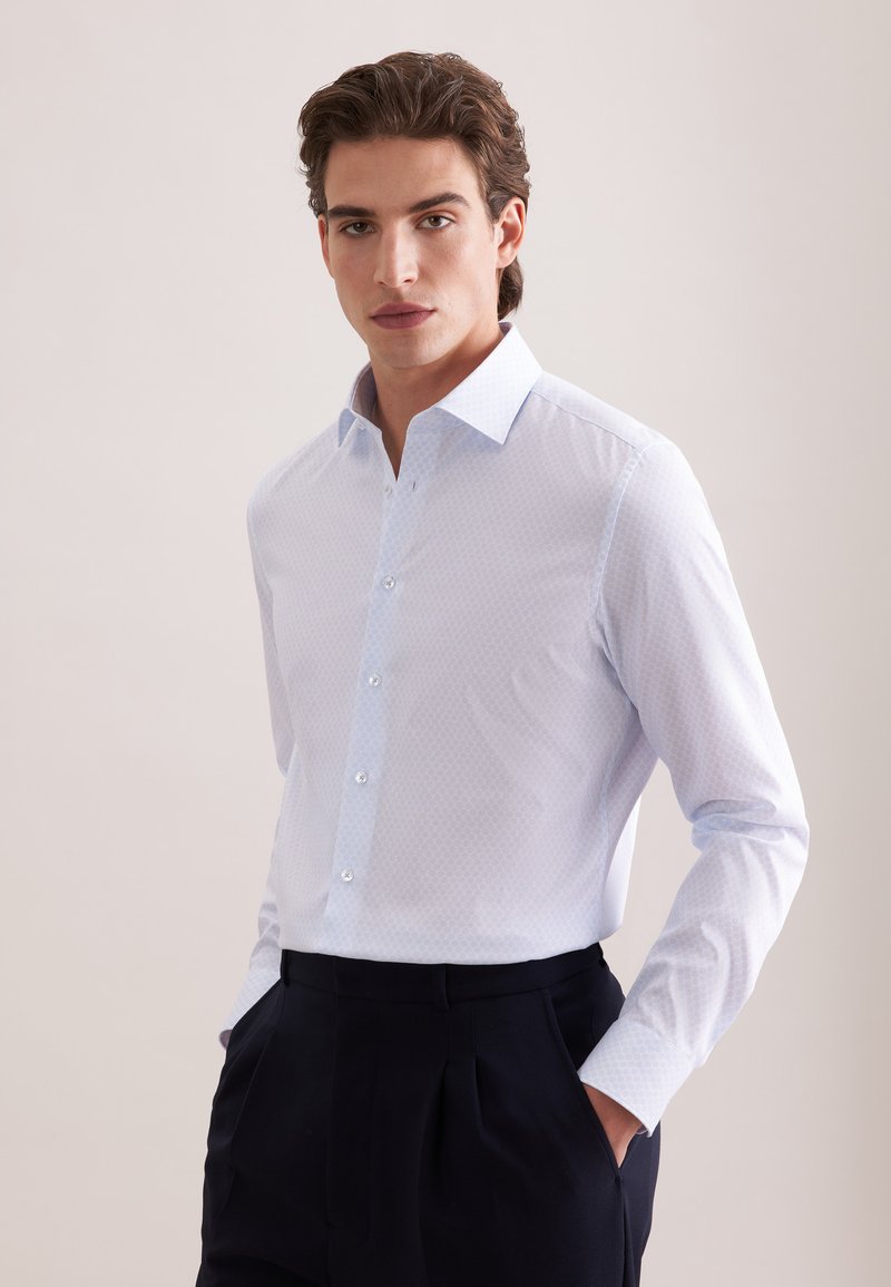 Jeune homme aux cheveux bruns coiffés, portant une chemise habillée bleu clair à motifs et un pantalon foncé, debout les mains dans les poches devant un fond neutre.