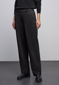 Pantalons noirs taille haute avec une finition texturée, présentant un design à jambes larges, des plis à l'avant et deux poches avant. Stylés avec des chaussures noires.