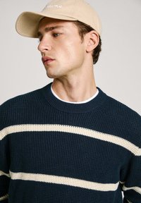 Maglione lavorato a maglia blu navy con strisce crema, scollo a coste e finitura testurizzata. Cappello beige con la scritta "Dreams" ricamata in bianco sulla parte anteriore.