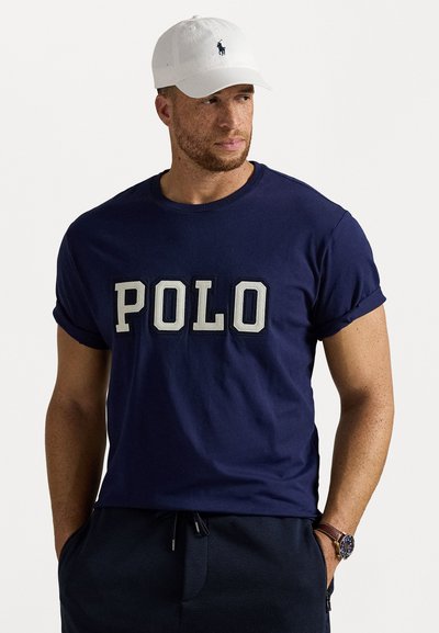 Granatowa bawełniana koszulka z napisem "POLO" w kremowym kolorze, okrągły dekolt, podwinięte rękawy; zestawiona z białą czapką z logo.