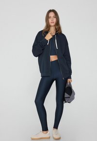 Marinefarbener Reißverschluss-Hoodie mit weißen Kordeln über einem passenden Tanktop und glänzenden Leggings. Helle Sneakers und eine graue Mütze in der Hand.