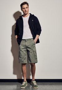 Černá zipová mikina, bílý tričko, olivově zelené cargo šortky a zelené tenisky s bílou podrážkou; vystavené na neutrálním pozadí.