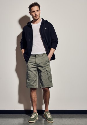 Schwarze Zip-Jacke, weißes T-Shirt, olivgrüne Cargo-Shorts und grüne Turnschuhe mit weißen Sohlen; vor einem neutralen Hintergrund präsentiert.
