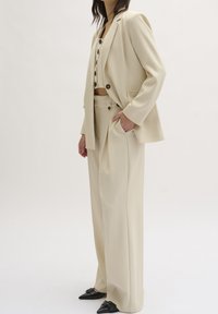 Traje ajustado color crema que presenta un blazer estructurado con botones negros, pantalones de cintura alta a juego y zapatos negros de punta.