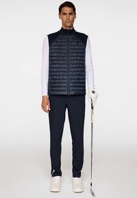 Marinblå vadderad väst med horisontellt quiltat mönster, dragkedja framtill, över en vit långärmad skjorta. Mörka byxor och golfklubba ingår.