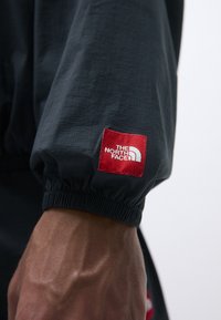 Manche de veste noire avec poignet froncé, ornée d'un patch logo rouge et blanc, mettant en valeur un tissu texturé.