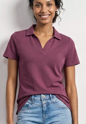 Smilende kvinde iført en kortærmet lilla skjorte med krave, stoppet ned i lyseblå jeans med høj talje, der står mod en hvid væg.