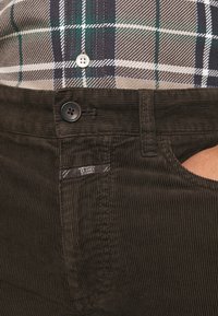 Pantalon en velours côtelé marron foncé avec une texture côtelée subtile, doté d'une fermeture par bouton et d'une étiquette à la taille. Porté avec une chemise à carreaux.