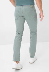 Grüne Slim-Fit-Jeans mit strukturiertem Gewebe, fünf Taschen, und einem Logo-Patch am hinteren Bund, kombiniert mit weißen Sneakers.