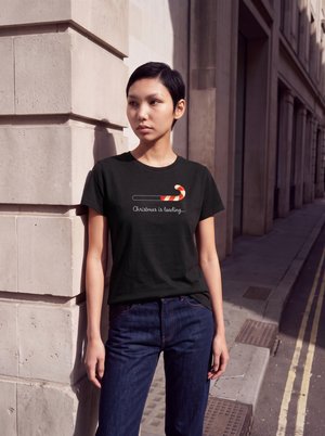 Una mujer está de pie junto a un edificio de piedra en una calle de la ciudad. Lleva una camiseta negra con un bastón de caramelo y las palabras «La Navidad se está cargando...»