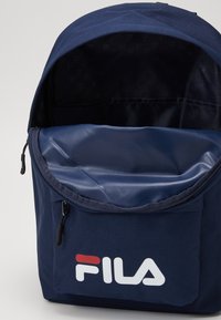 Zaino in tessuto blu con chiusura a zip, ampio scomparto principale e tasca frontale con logo Fila bianco. Rivestimento interno testurizzato.