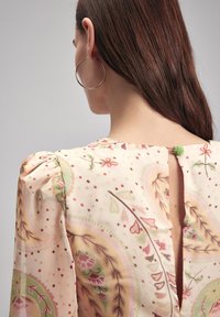 Bluse mit floralen Mustern in Pastellfarben, mit Puffärmeln und einem Schlüssellochverschluss am Rücken, der mit einem dekorativen grünen Knopf versehen ist.