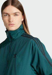 Teal zip-up jakke lavet af let stof. Har en høj krave, sort Adidas-logo og tre sorte striber på hver skulder.
