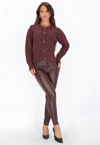 Burgundská pletená cardigan s diamantovými vzory a stříbrnými akcenty, zapínání na knoflíky, spárovaná s burgundskými legínami z umělé kůže a černými podpatky.