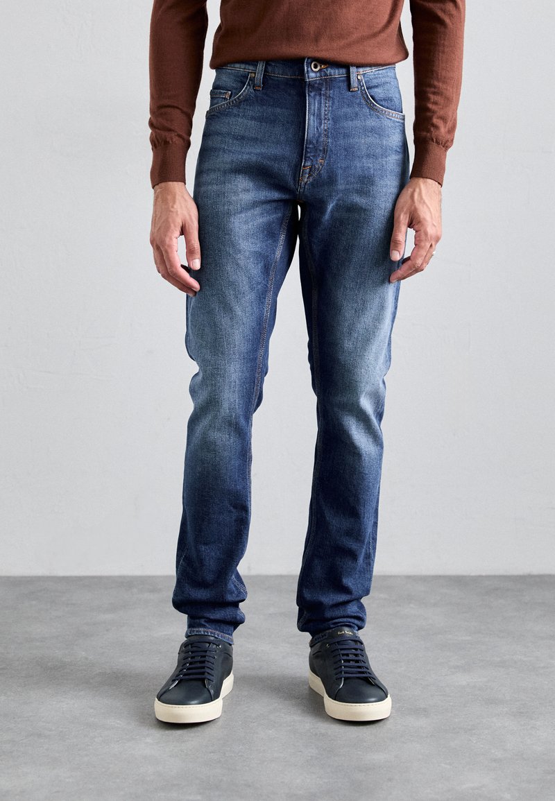 Jeans de denim azul com um corte slim, apresentando detalhes desgastados, fecho com botão e ziper, e design de cinco bolsos. Combinados com ténis escuros.