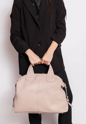 Persoon draagt een zwarte blazer en houdt een grote lichtbeige leren handtas vast bij beide handvatten voor een witte achtergrond.